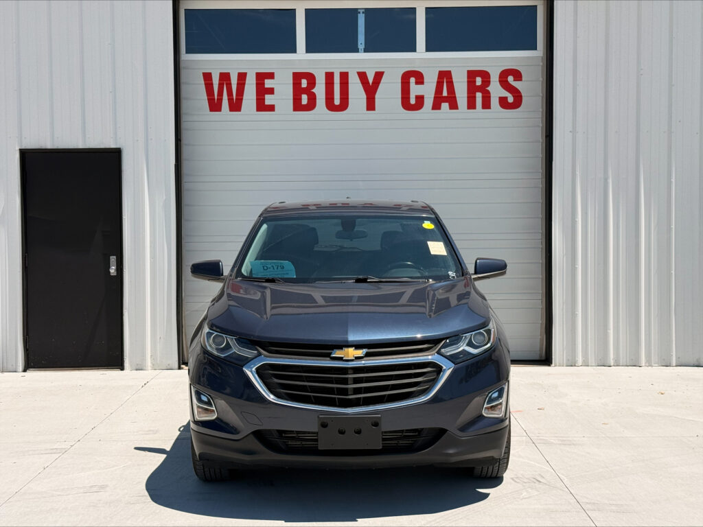 Car parked in front of a white garage door with red 'WE BUY CARS' sign above it, front view of dark Chevrolet SUV.