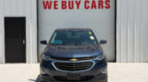 Car parked in front of a white garage door with red 'WE BUY CARS' sign above it, front view of dark Chevrolet SUV.