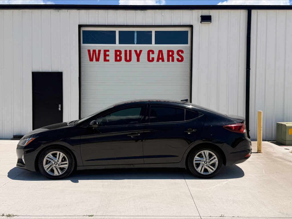 Black sedan parked in front of a white building with a garage door that reads 'WE BUY CARS' in red letters.