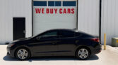 Black sedan parked in front of a white building with a garage door that reads 'WE BUY CARS' in red letters.