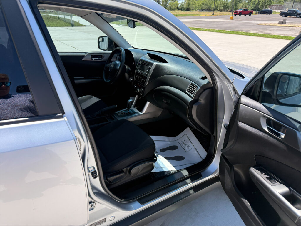 Interior of a silver SUV with the driver's seat, steering wheel, and center console visible through an open front door in a parking lot, with a person’s reflection in the window.