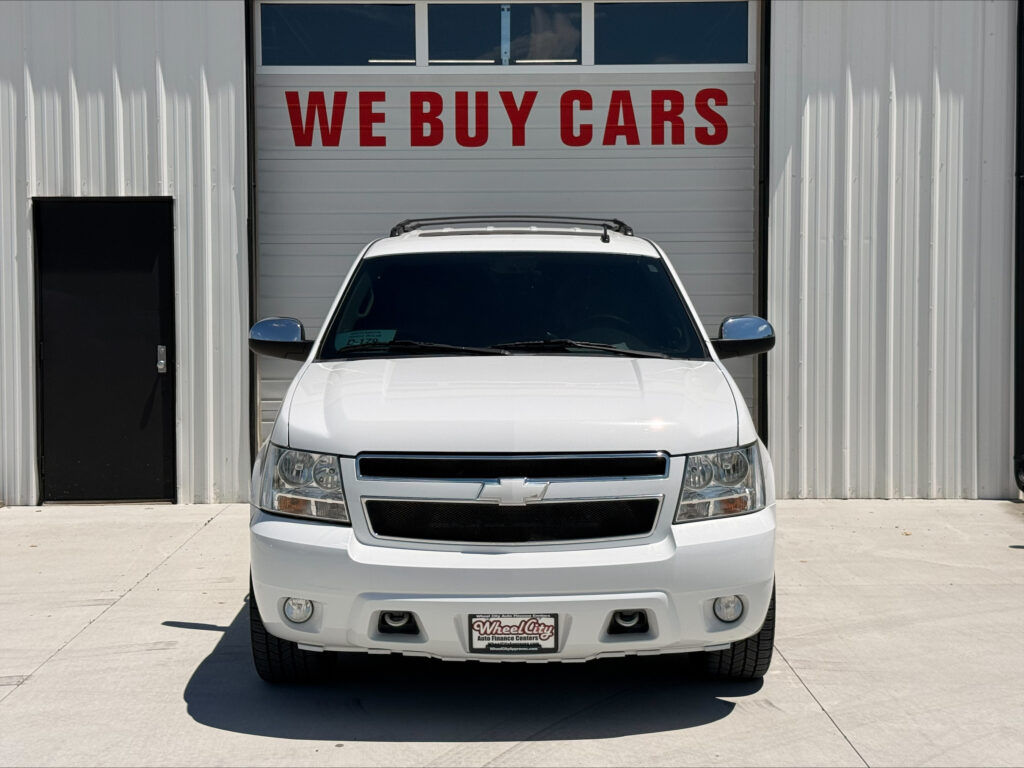 White SUV facing the camera in front of a building with a large red 'WE BUY CARS' sign above it