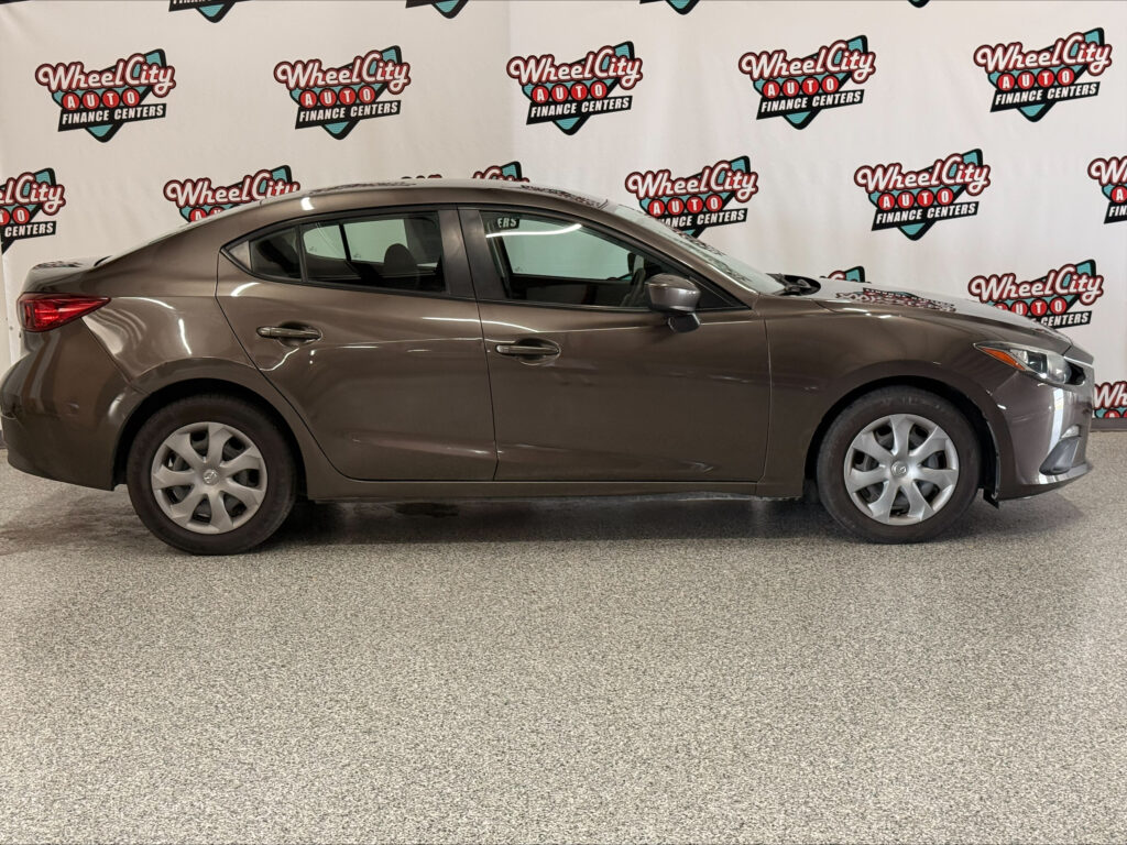 Brown four-door sedan indoors in a showroom with Wheel City Auto Finance Centers logos on a white backdrop behind it.