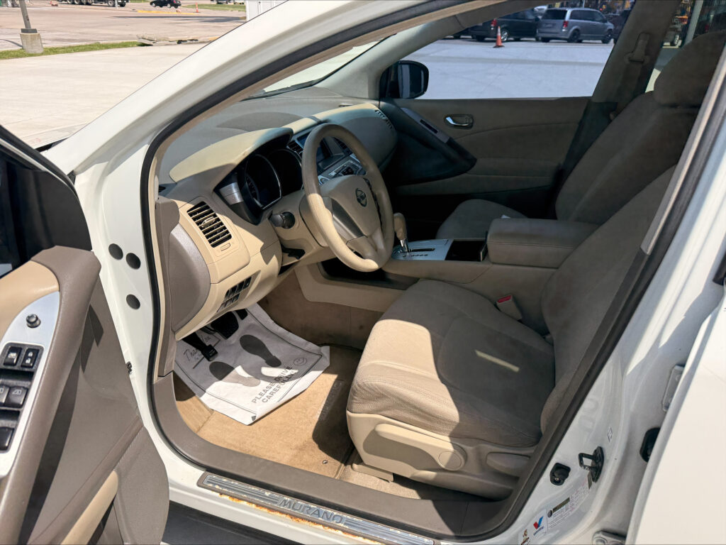 Open driver's door view of a beige SUV interior showing the steering wheel, dashboard, center console, and front seats; a parking lot is visible outside the windows.