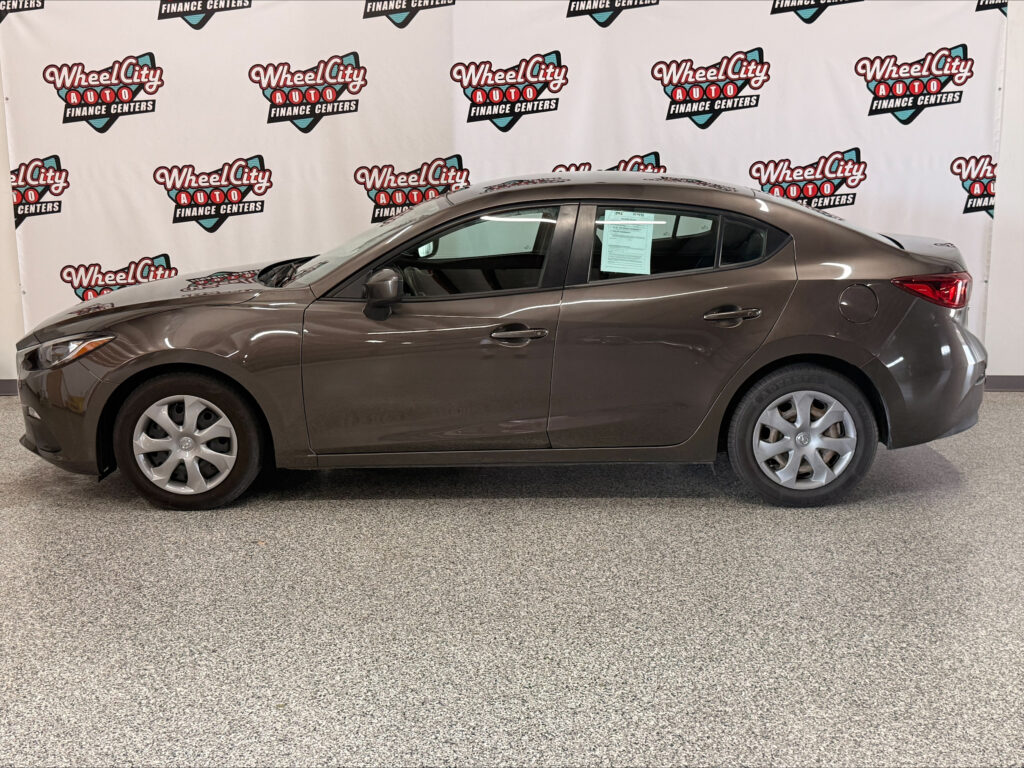 Brown mid-size sedan photographed indoors in front of a Wheel City Auto Finance Centers backdrop, shown from the passenger side with visible windows, door handles, and rear taillight.