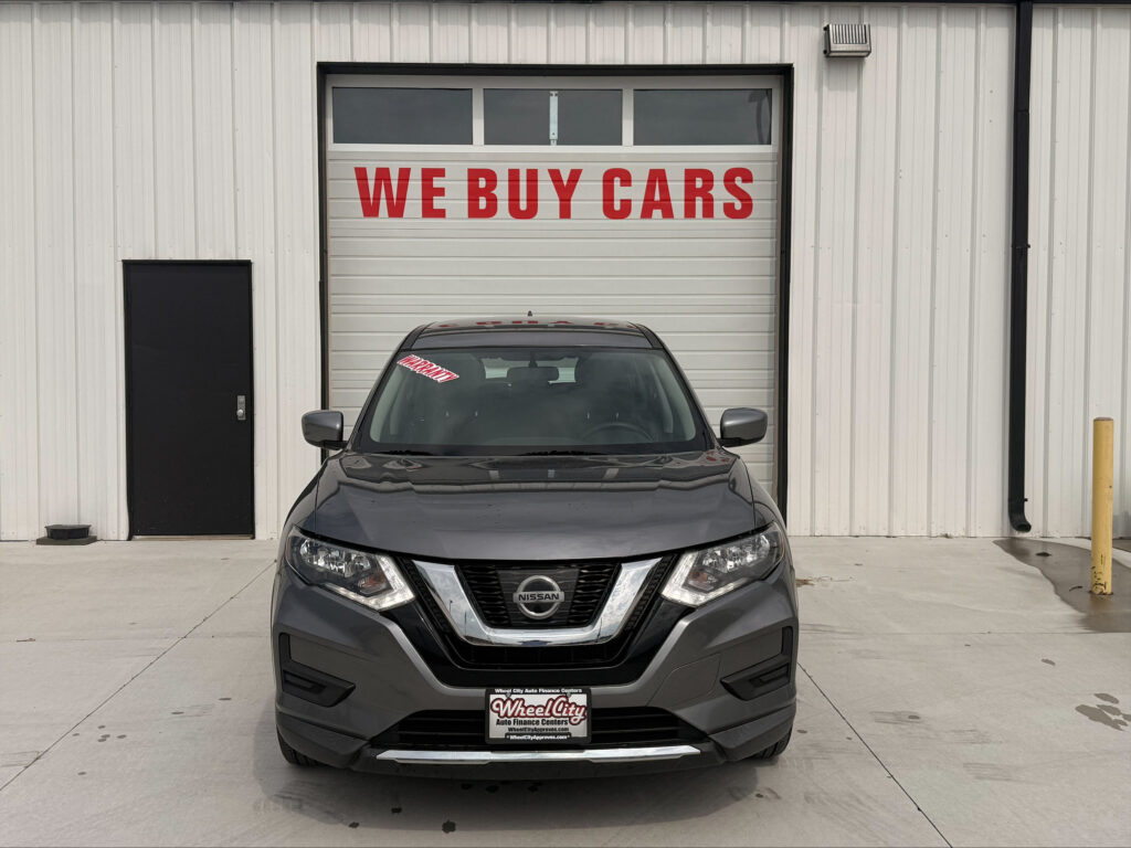 Gray Nissan SUV parked in front of a white industrial building with a large garage door that reads 'WE BUY CARS' in red letters above the car's windshield window.
