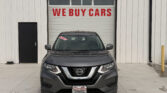 Gray Nissan SUV parked in front of a white industrial building with a large garage door that reads 'WE BUY CARS' in red letters above the car's windshield window.