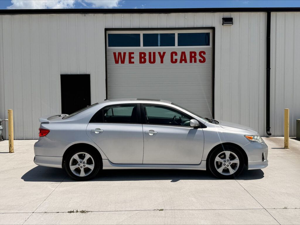 Silver sedan parked in front of a white industrial building with a large garage door that reads 'WE BUY CARS' in red letters outside.