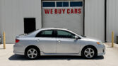 Silver sedan parked in front of a white industrial building with a large garage door that reads 'WE BUY CARS' in red letters outside.