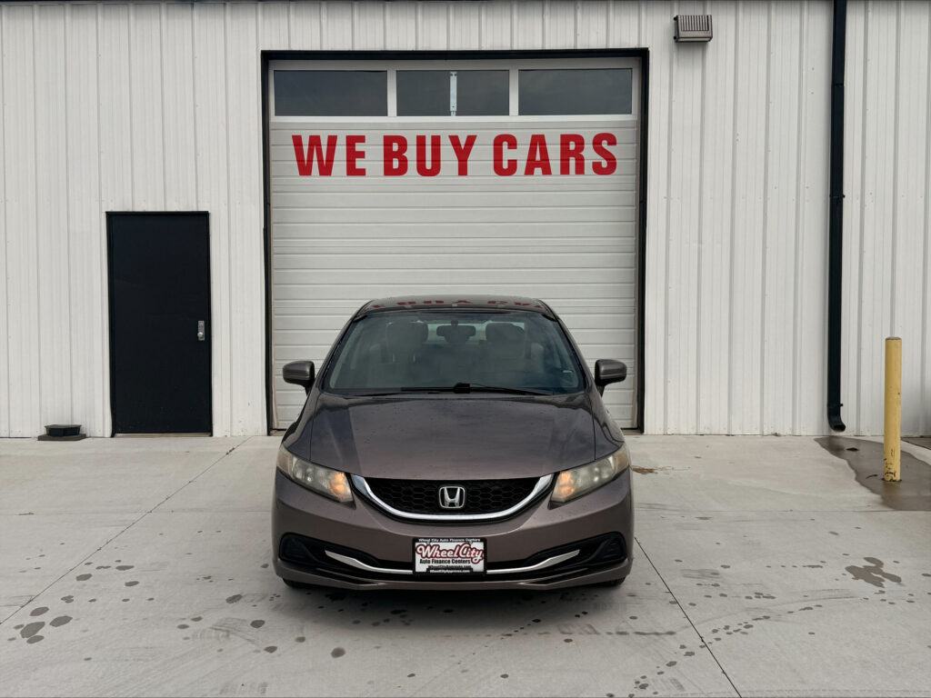 Front view of a gray Honda parked in front of a white garage door with 'WE BUY CARS' in red letters loping across the door.