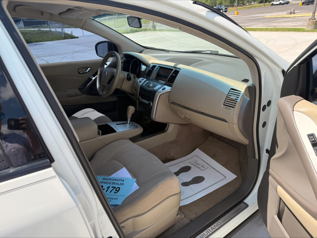 Interior of a white Nissan Murano with beige seats and center console visible, driver’s-side door open and dashboard in view; dealership floor mats on the front passenger side.