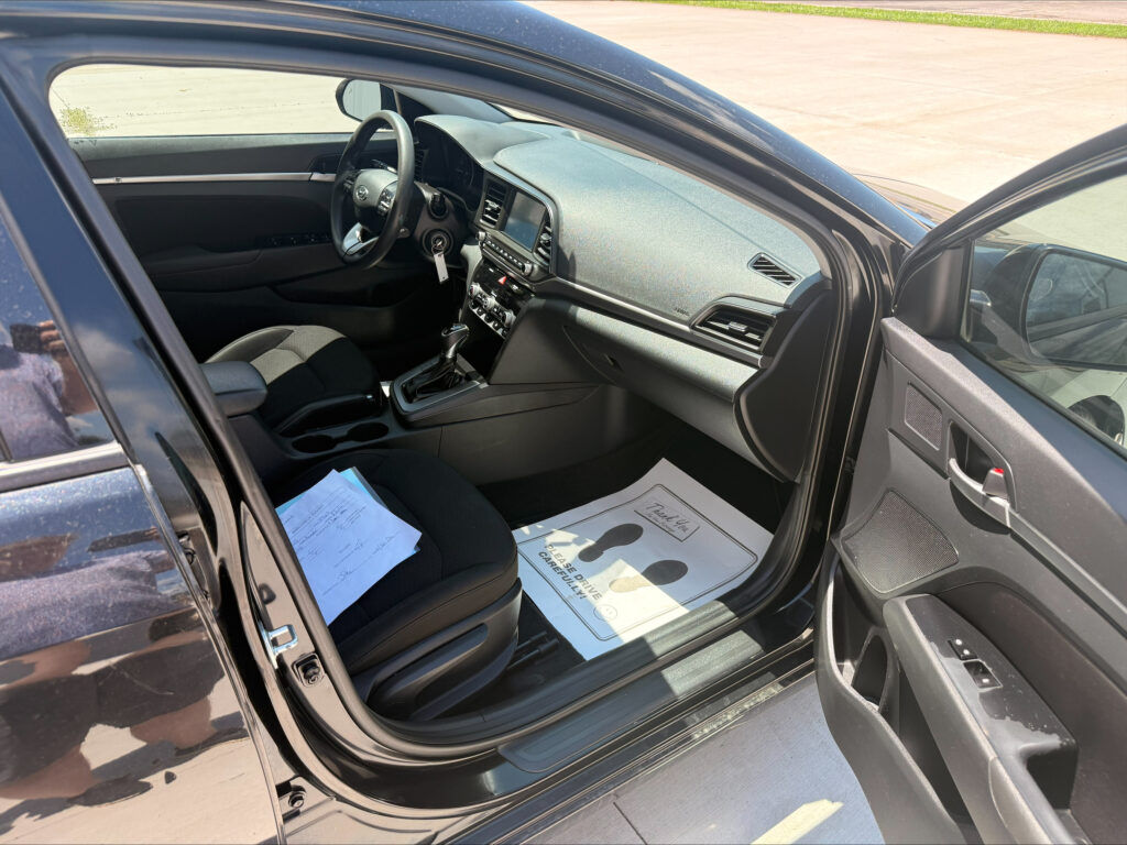 Interior of a dark gray car with the driver’s seat, steering wheel, center console and dashboard visible; papers lie on the front passenger seat and the door is open on the left side.