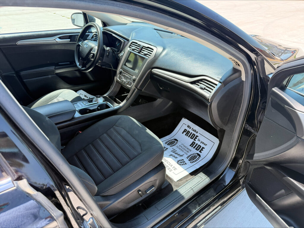 Interior of a car shows driver and passenger seats, steering wheel, center console, and paper floor mat visible on the passenger side