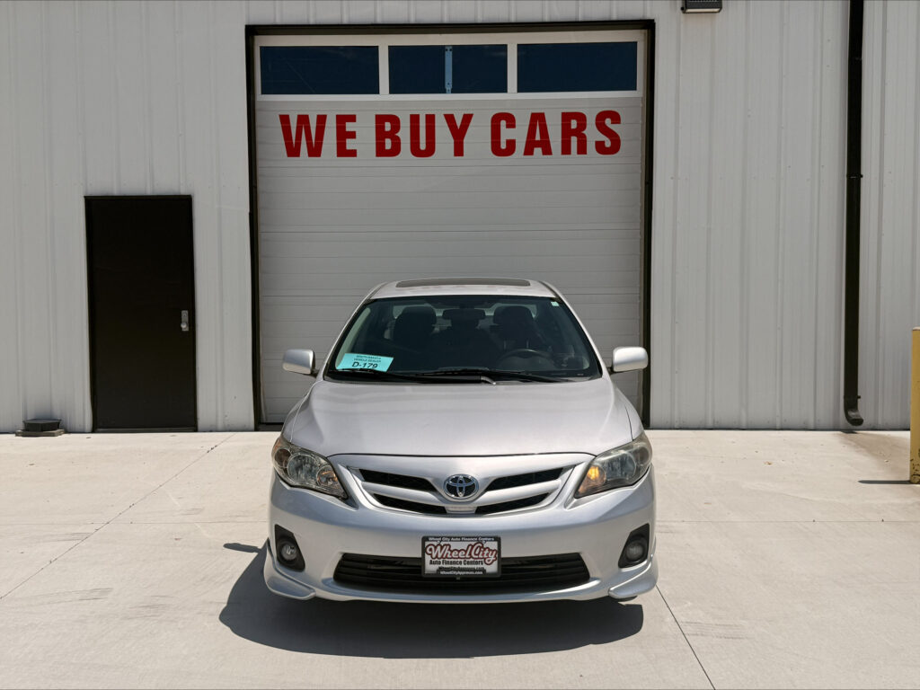 Front view of a silver Toyota car parked in front of a garage door with a bold red 'WE BUY CARS' sign above it.