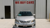 Front view of a silver Toyota car parked in front of a garage door with a bold red 'WE BUY CARS' sign above it.