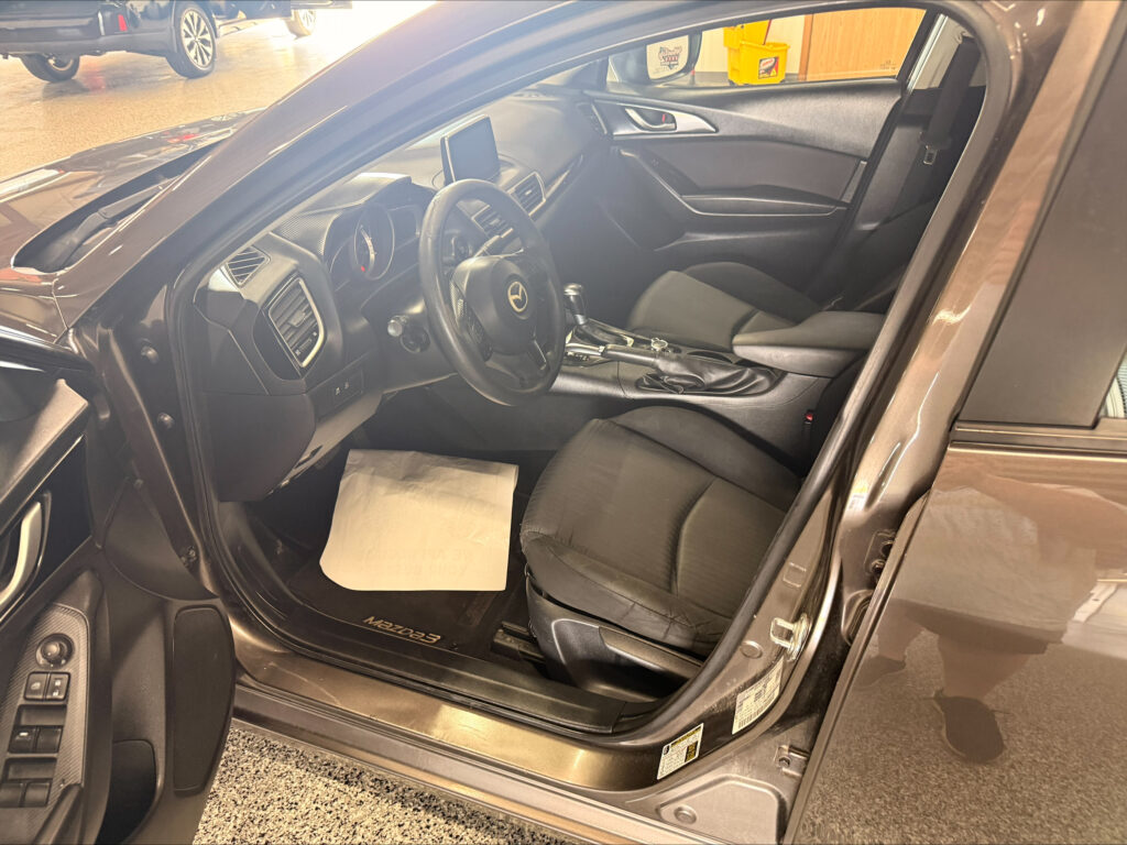 Interior of a Mazda3 with the driver's door open, showing the steering wheel, center console, and black fabric seats in a showroom.
