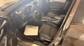 Interior of a Mazda3 with the driver's door open, showing the steering wheel, center console, and black fabric seats in a showroom.