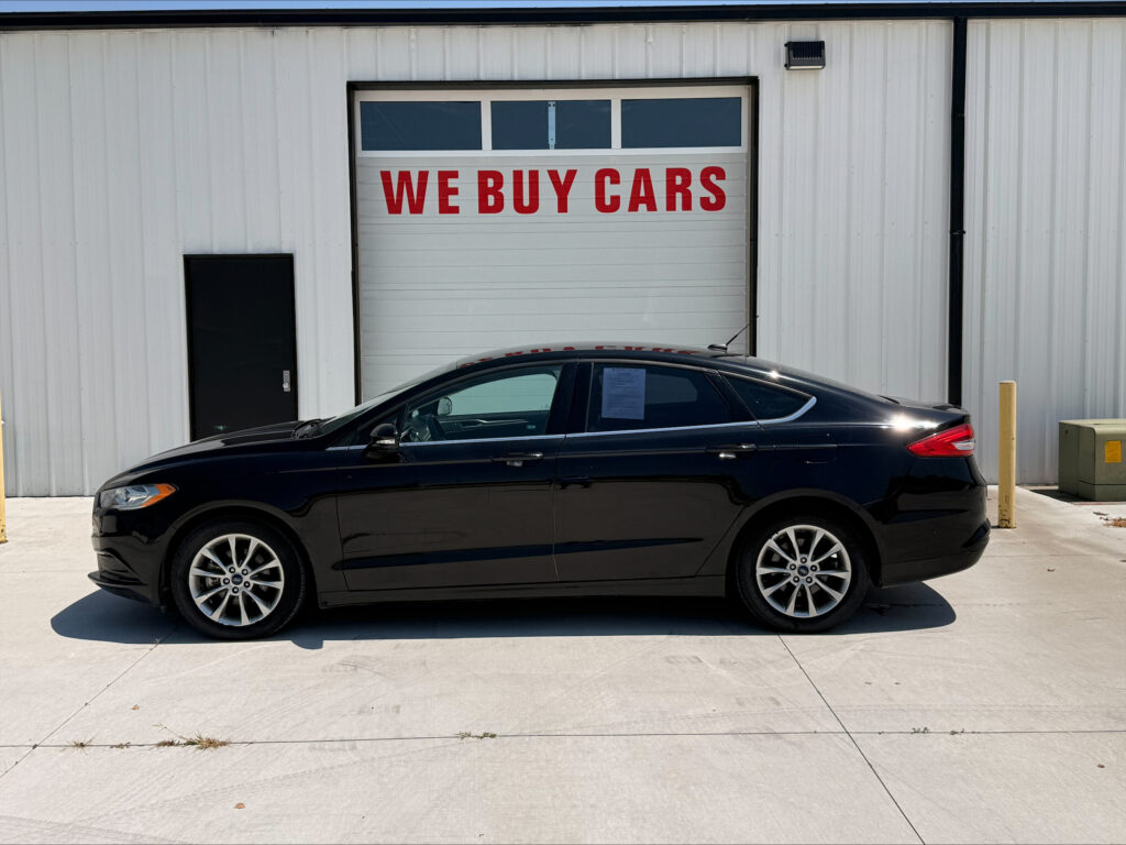 Black sedan parked in front of a white industrial building with a large door that reads 'WE BUY CARS' in red letters above the windowed section.