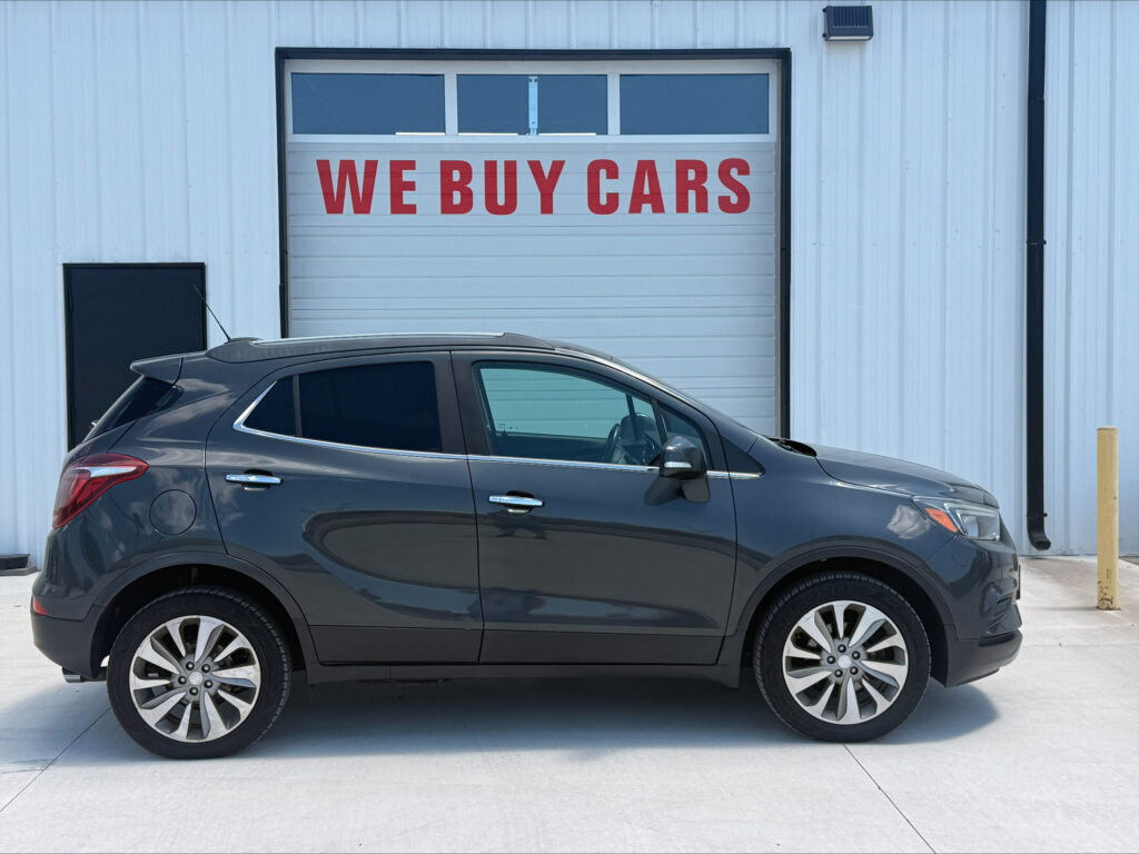 Gray SUV parked in front of a white industrial building with a garage door that reads 'WE BUY CARS'.