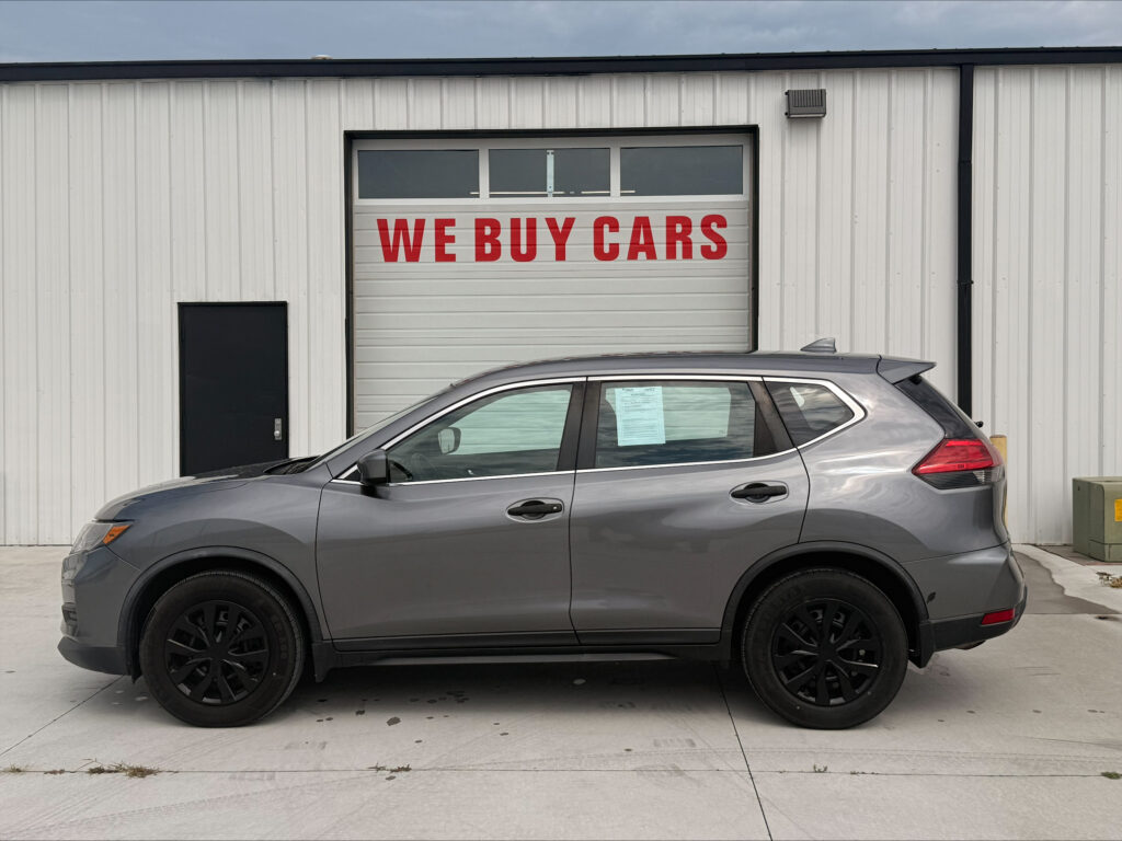 Gray SUV parked in front of a white corrugated metal building with a large red 'WE BUY CARS' sign on the garage door.