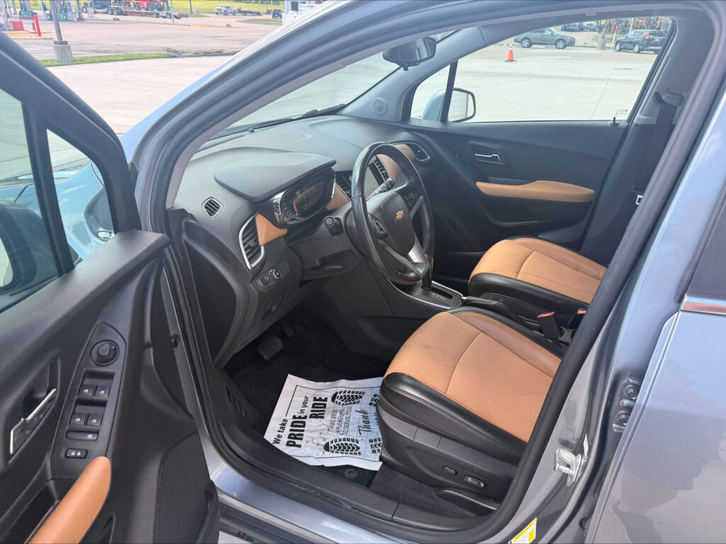 Interior view of a car with the driver's seat and steering wheel visible, tan leather seats, and the front passenger door open; a dealership floor mat is on the floor and the parking lot is visible outside the window.