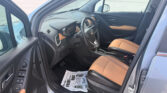 Interior view of a car with the driver's seat and steering wheel visible, tan leather seats, and the front passenger door open; a dealership floor mat is on the floor and the parking lot is visible outside the window.