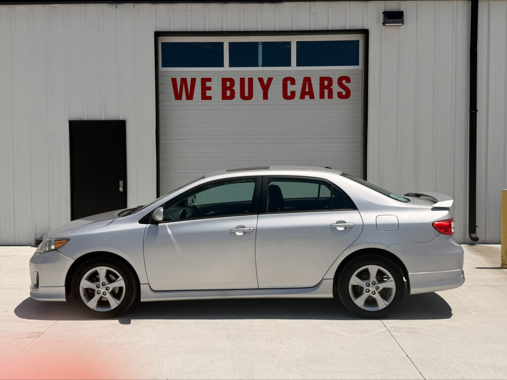 Silver sedan parked in front of a white industrial building with a large garage door that says 'WE BUY CARS' in red letters