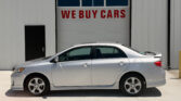 Silver sedan parked in front of a white industrial building with a large garage door that says 'WE BUY CARS' in red letters
