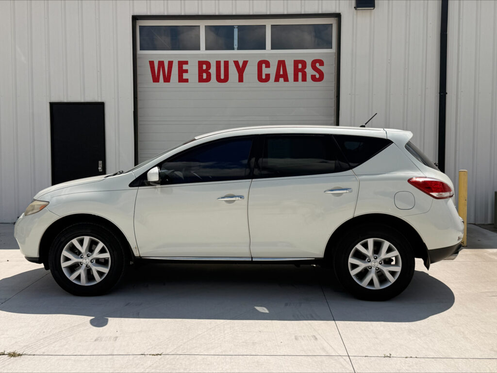 White SUV parked in front of a gray industrial building with a large garage door reading WE BUY CARS.