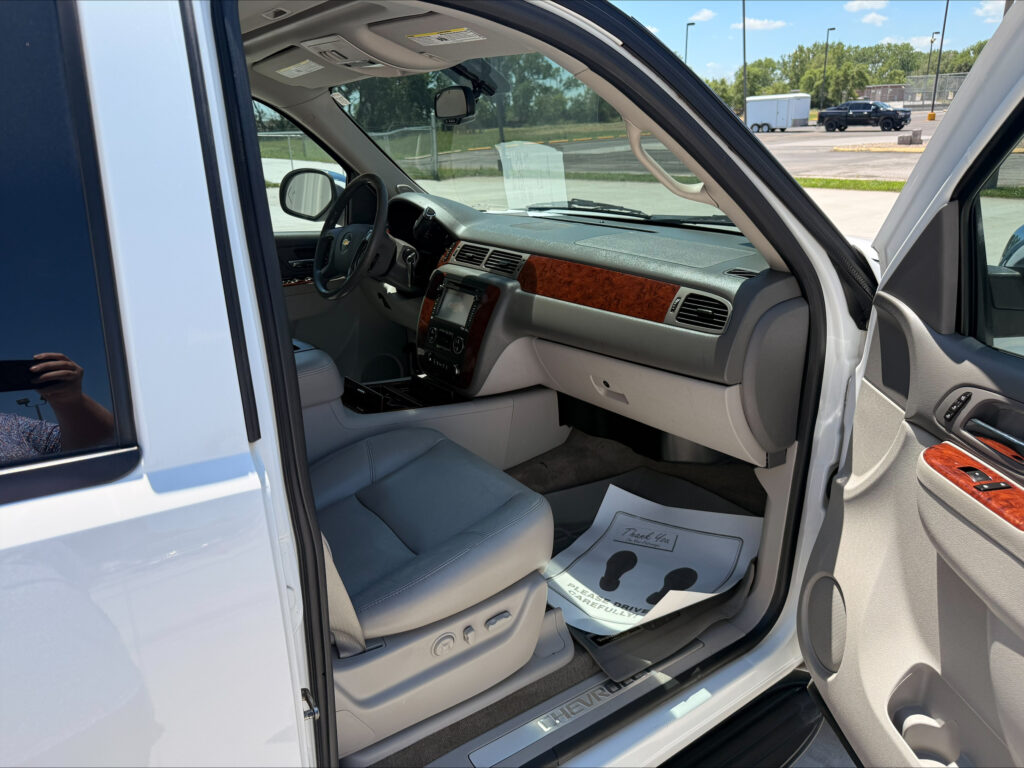 Open driver's door of a white Chevrolet SUV, showing gray leather seats, wood-grain dashboard, and built-in screen in the center console; parking lot visible outside.