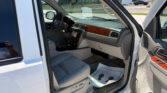 Open driver's door of a white Chevrolet SUV, showing gray leather seats, wood-grain dashboard, and built-in screen in the center console; parking lot visible outside.