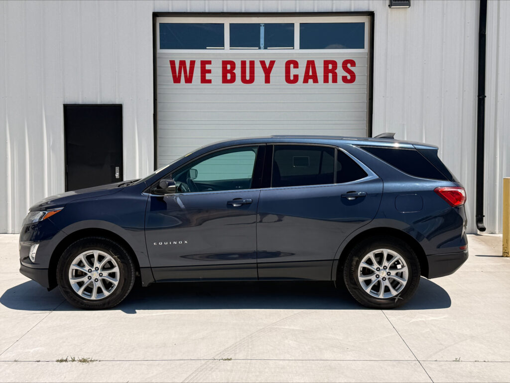 Blue Chevrolet Equinox parked in front of a white industrial building with a garage door reading WE BUY CARS in red letters beneath a windowed panel area to the left of the door side.