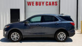 Blue Chevrolet Equinox parked in front of a white industrial building with a garage door reading WE BUY CARS in red letters beneath a windowed panel area to the left of the door side.