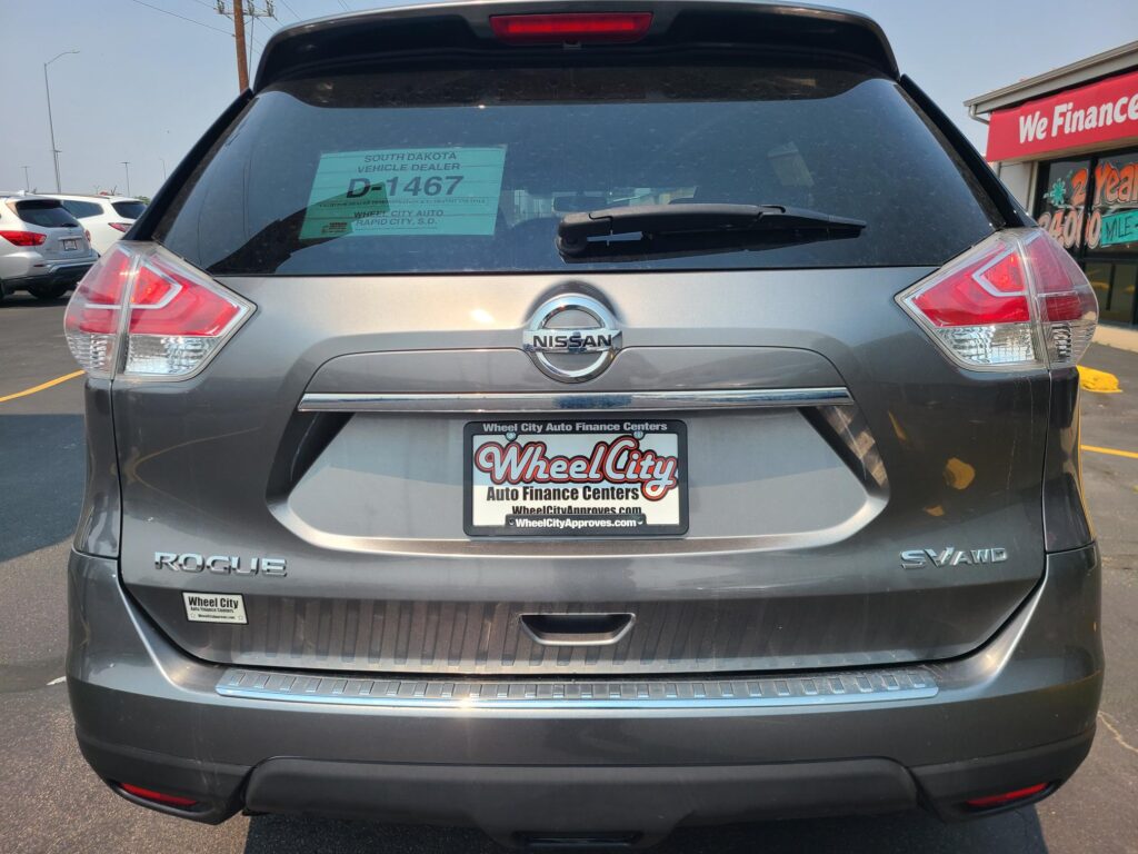 Rear view of a gray Nissan Rogue SUV with a Wheel City Auto Finance Centers plate frame in a parking lot; green dealership sticker on rear window and a storefront sign in the background: 'We Finance'