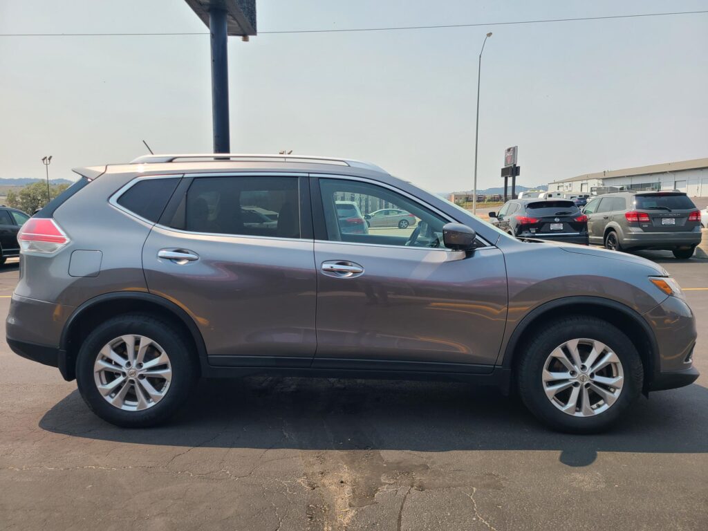 Side view of a silver SUV with roof rails parked in a lot, other cars and an industrial building visible in the background
