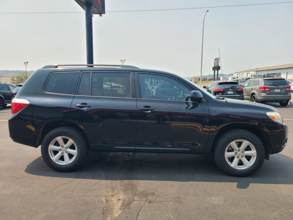 Side view of a dark SUV with roof rails parked in a busy dealership lot, other cars and a building visible in the background.