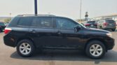Side view of a dark SUV with roof rails parked in a busy dealership lot, other cars and a building visible in the background.