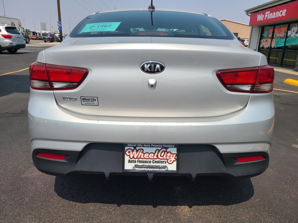 Rear view of a silver Kia Rio in a dealership lot, with Wheel City license plate frame and a 'We Finance' storefront in the background.