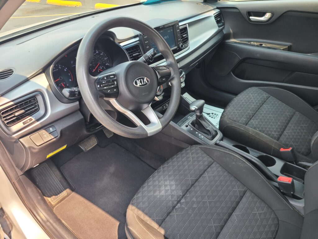 Front interior of a Kia: steering wheel, instrument cluster, center touchscreen, gear shifter, and black cloth seats in view.