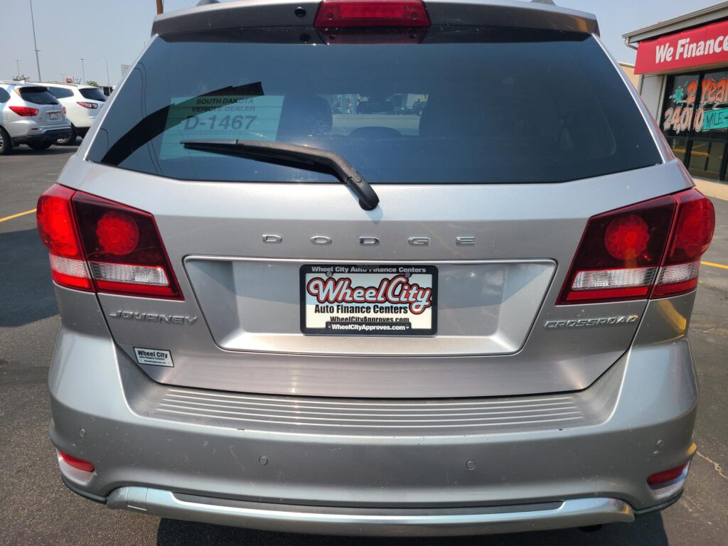 Rear view of a silver Dodge Journey in a car dealership lot, with a D-1467 window sticker and Wheel City plate frame visible on the license plate area.