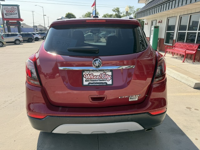 Rear view of a red Buick Encore SUV parked in a dealership lot; Buick emblem on the tailgate, 'Encore' badge visible, license plate frame reads 'Wheel City Auto Finance Centers' with surrounding dealership signage in the background.