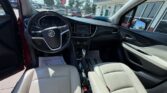 Interior of a Buick car showing beige leather seats, steering wheel with Buick emblem, center console and gear shifter; front doors open, dealership lot visible through the windshield.