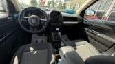 Interior of a Jeep SUV: view from the front seats showing steering wheel, center console with radio and climate controls, gear shifter, and front passenger seat; outside is a parking lot visible through windows.