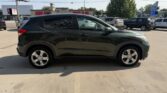 Side view of a dark green compact crossover SUV parked in a dealership lot with other cars and storefronts in the background.