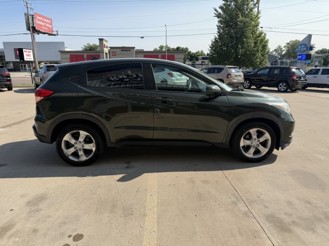 Side view of a dark green compact crossover SUV parked in a dealership lot with other cars and storefronts in the background.