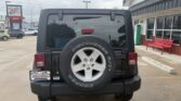 Rear view of a black Jeep Wrangler with a mounted spare tire on the tailgate, parked in a dealership lot near a red bench and building.