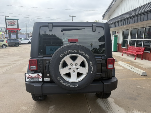 Rear view of a black Jeep Wrangler with a mounted spare tire on the tailgate, parked in a dealership lot near a red bench and building.