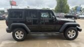 Black four-door Jeep Wrangler with a hardtop parked in a dealership lot, side profile visible with large tires and black exterior