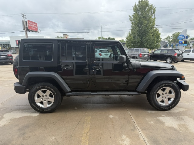 Black four-door Jeep Wrangler with a hardtop parked in a dealership lot, side profile visible with large tires and black exterior
