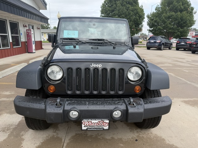 Front view of a black Jeep Wrangler with a seven-slot grille, round headlights, and a rugged bumper on a wet parking lot; dealership plate frame reads Wheel City Auto Finance Centers.